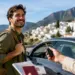 Turista junto a un coche mientras recibe una llave, con documentos de viaje y un pueblo costero de fondo