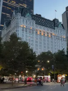 The Plaza hotel in New York at dusk, with trees, traffic and street vendors in front